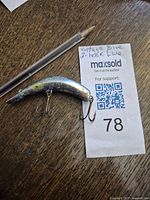 Photo showing vintage blue fishing lure with two treble hooks placed on a wooden surface next to a pen and a MaxSold label.