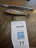 Photo showing the vintage fishing lure beside a pen for scale, displaying its black-grey coloring and metal hooks.