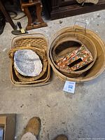 View of two groups of baskets on a concrete floor, showing the variety of sizes and shapes including wicker and wooden baskets and inset mosaic pattern basket.
