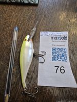 Photo of the yellow-white vintage fishing lure next to a pen for scale, showing its shape, color, and two treble hooks attached.