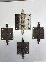 Four antique Victorian-style metal door hinges on a white surface; three smaller 3-inch hinges with decorative finials and one larger 4-inch hinge with simpler design and visible aging