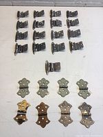 Top-down view of 17 smaller decorative cabinet hinges and 8 larger plain cabinet hinges laid flat showing wear and paint marks