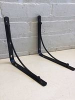 Pair of black metal L-shaped shelf brackets with curved support arms, standing vertically against a light gray brick wall, showing front and side views.