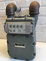 Front view of the Canadian Meter Company Limited gas meter showing mechanical dial display marked 'Cubic Feet' and model plate 'TYPE ALC 175'. Two rusted L-shaped metal pipes attached on top.