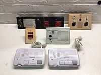 Full view of all items including intercom units and nurse call device panels arranged on a white surface against grey brick background.
