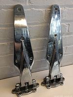 Pair of large, polished metal finish hinges standing upright against a textured wall, showing size and shape.
