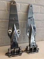 Front view of two large polished metal hinges standing upright showing their shape and screw holes.