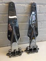 Photo showing two tall, pointed vintage fridge hinges standing upright on a surface against a white brick wall background.