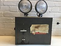 Front view of large grey metal emergency light with two circular adjustable lamps, control knobs and buttons, and a worn paper label.