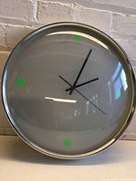 Front view of round wall clock showing white face, black hands, silver metal frame, and green markers at 12,3,6,9 positions.