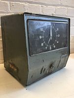 Front and side angle of vintage mechanical time clock showing circular dial and metal casing with wear marks.