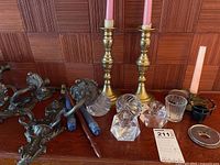 Photo showing brass candle holders, wall sconces, glass candle snuffers and other small candle accessories on a wooden surface with wood panel background.