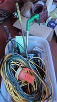 Photo showing various electrical cables and cords tangled together including yellow extension cords along with gardening tools and a plastic tote beneath.