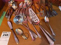 Top view of assorted silver plated serving spoons, forks, knives, and serving pieces on a table with a Maxsold lot tag visible. Some items show tarnish and varied handle designs.
