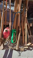 Multiple wooden-handled gardening tools including rakes, shovels, and hoes leaning against a wall, with a red watering can and a green handled tool in front.