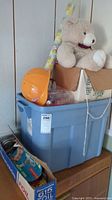 Blue storage bin containing toys with a beige teddy bear sitting on top and decorative paper roll visible.