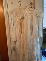 Two vintage dresses hanging on a door, one in a protective plastic cover, showing full length with lace and chiffon fabric.