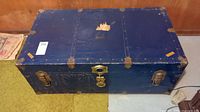 Vintage blue metal trunk closed showing top and front side with metal latches and corner protectors, visible wear and tear.