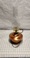 Antique copper hurricane lamp with clear glass chimney, electric cord attached, shown unlit on grid background