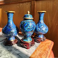 Three cloisonné pieces and stands arranged on marble-top table in sunlight