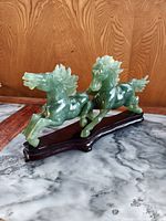 Pair of green hard stone horses carved in running pose on a dark wooden stand placed on marble surface, front side view.