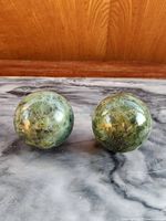 Two green polished stone spheres displayed side by side on a grey marble surface with wood backdrop, showing natural mottled green shades and surface texture.