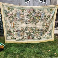 Full front view of the large silk tapestry held outdoors showing full detailed village scene with trees, buildings, and people, including fringed edges