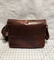 Front view of the brown leather messenger bag showing the flap and stitched edges on a grid-patterned background.