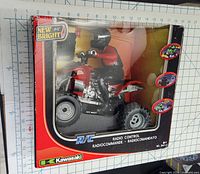 Front view of the Kawasaki remote controlled ATV in sealed box with grid background containing measurement markings.