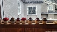 Eight ceramic mushrooms displayed on a wooden windowsill with a textured window background. Caps vary in red, dark purple, and muted green/tan colors; stems are white and uniform.