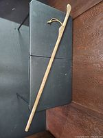 Full length view of the oak walking stick laid diagonally on a black folding table against a wooden wall background showing natural wood grain and curved handle.