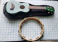 Top view showing green Makala ukulele on a black case, six strings visible, and wooden tambourine with metal jingles arranged around the frame with one jingle missing.