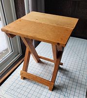 Light brown wooden plant stand with rectangular top and X-shaped collapsible legs seen from front left angle.