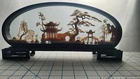 Full front view of oval glass case diorama on wooden base showing detailed cork carvings of Chinese buildings and trees.