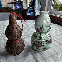Photo showing both double gourd shaped vessels side by side on a checkered tablecloth near a window with outdoor view, highlighting their contrasting materials and decorations.