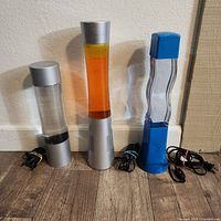 Three lava lamps displayed unlit against a wall, showing designs and colors of liquid and wax inside.