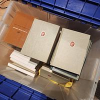 Photo showing multiple hardcover and softcover books in a plastic container, including two tan hardcover books and other smaller books