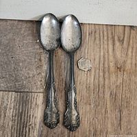 Two ornate sterling silver spoons and one small silver charm laid out on a wooden surface showing overall condition and design.