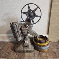 Front side of Keystone Regal film projector with seven-spoke film reel loaded and five film reel cases stacked next to it.