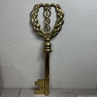 Front view of large solid brass key with laurel wreath and caduceus symbol at the bow, showing overall shape and details.
