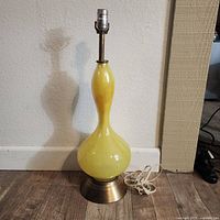 Full lamp with curvy yellow glass body and brass base on a wooden floor against a beige wall. The lamp has no bulb or shade attached.