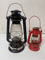 Photo showing both lanterns side by side: a larger black kerosene lantern and a smaller red one.