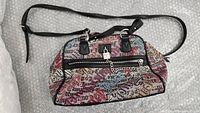Full view of Juicy Couture handbag with multi-logo print and black leather strap laid flat on white textured surface.