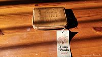Photo showing the brown Anna Paola wallet with attached branded tag, highlighting its leather crocodile texture and zip-around style.