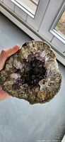 Hand holding a hollow oval geode showing purple amethyst quartz crystals inside surrounded by lighter quartz and rough outer stone.