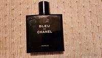Front view of Bleu De Chanel 100ml bottle on beige textured surface, showing gold lettering and black cap.