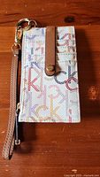 Front view of white leather Calvin Klein wallet with multicolor 'ck' print and tan leather snap closure tab attached.