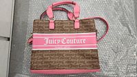 Full view of Juicy Couture handbag showing brown fabric with repeating logo pattern, pink leather handles, pink leather base and side accents, and a large pink horizontal stripe with Juicy Couture branding.