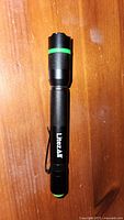 Full view of black LitezAll LED flashlight with green accent rings laying on wooden surface, showing clip side and textured grip body.