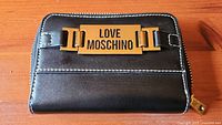 Front view showing black leather wallet with gold 'LOVE MOSCHINO' logo plate and stitching detail.